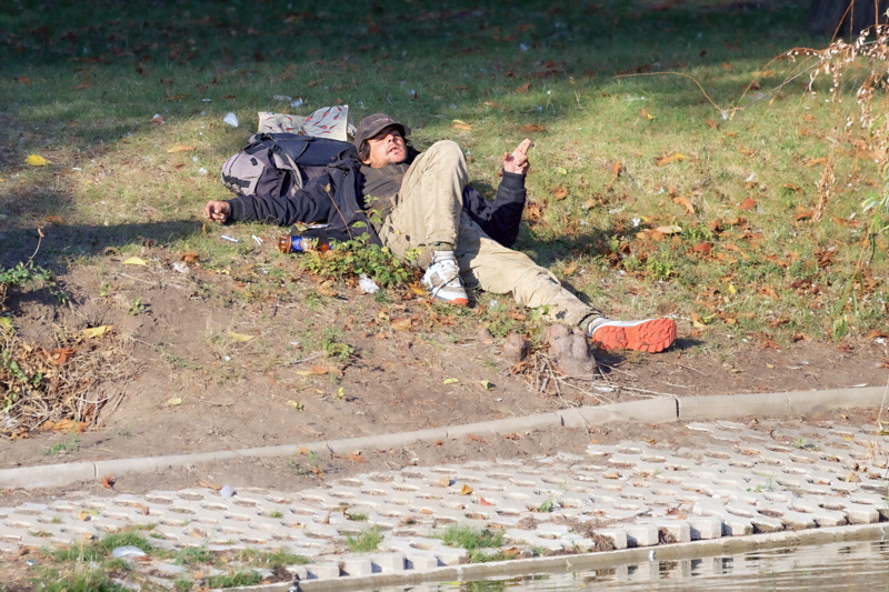 16 - A man lying down on a grassy slope outdoors next to a paved embankment and water, wearing a dark cap, jacket, and light trousers, with a backpack and a beverage container nearby